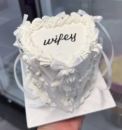 A white heart shaped mini cake with ruffled piping and white ribbons, written wifey